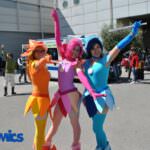 Tre cosplayer colorati in pose festose al ROMICS di Roma, durante un evento di fumetti e cultura pop, circondati da un vivace pubblico.