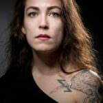 Ritratto di una donna con capelli scuri e occhi chiari, che mostra un tatuaggio di un uccello, in un contesto evocativo e intenso.