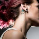 Immagine di profilo di una giovane donna con capelli rosso porpora e un tatuaggio di un quadrifoglio sul collo, simbolo di speranza e ricordo.