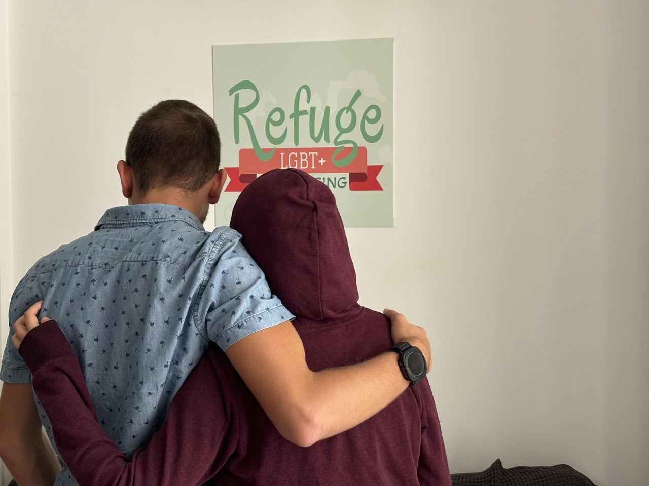 Due persone abbracciate davanti a un cartello che promuove un rifugio per comunità LGBTIQ+, simbolo di accoglienza e solidarietà.