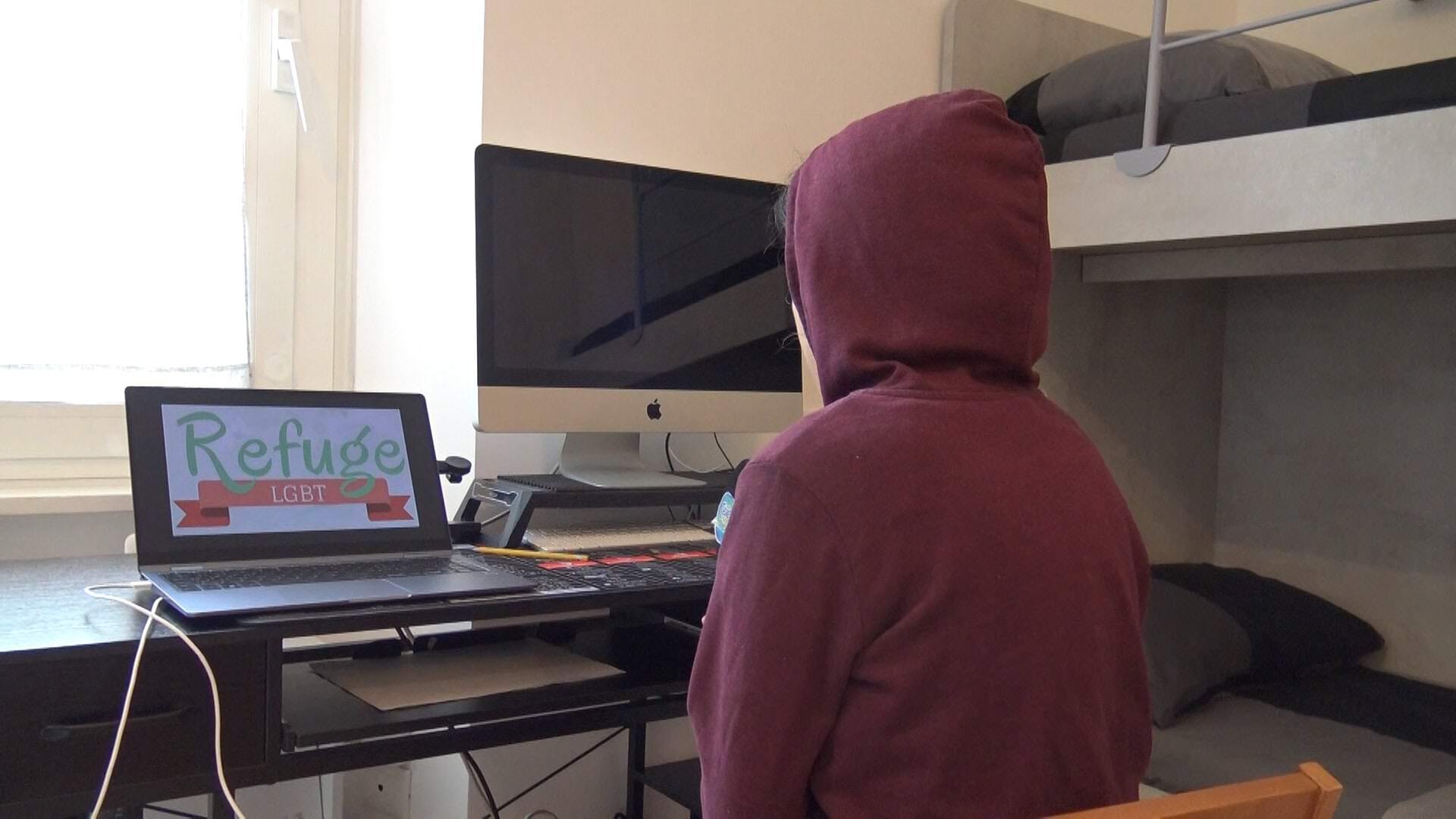 Una persona seduta di spalle in una stanza, davanti a un computer, esplora risorse online su rifugi per la comunità LGBT+.