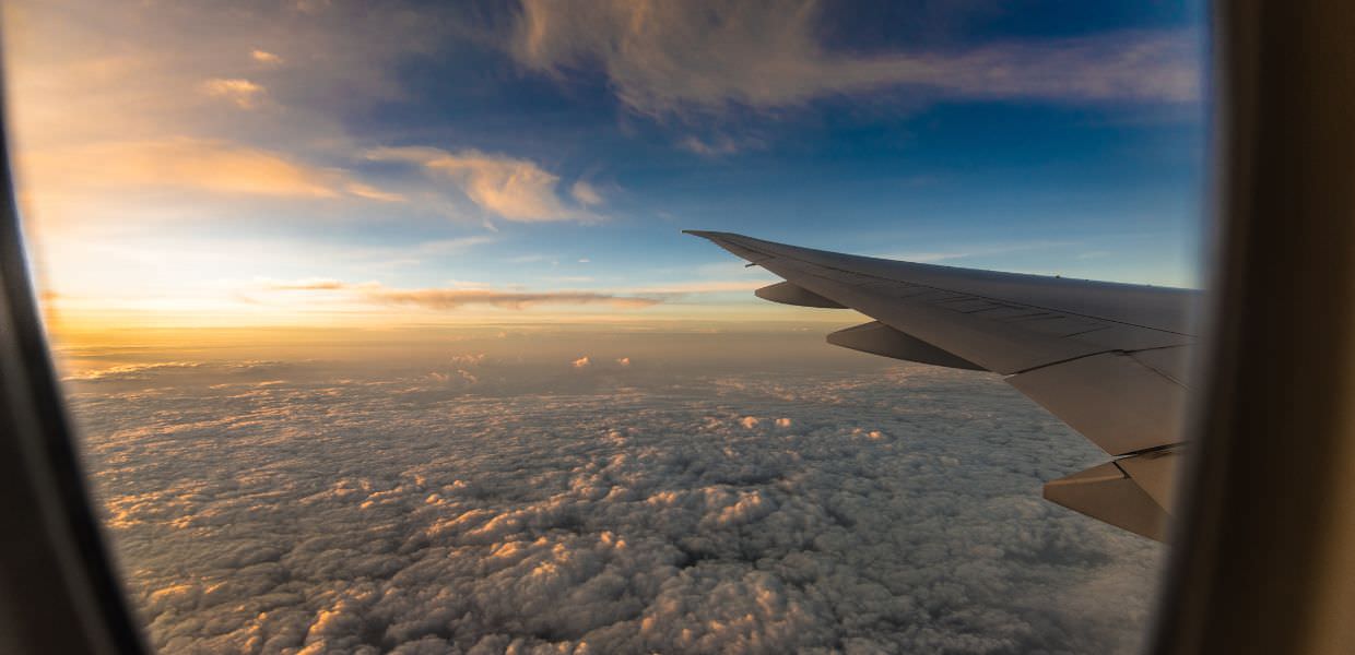 Vista aerea dal finestrino di un aereo, con nuvole sottostanti e un cielo al tramonto, illustrando il tema della crisi climatica e dei viaggi.