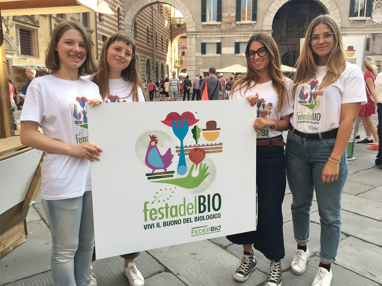 Quattro giovani sorridenti presentano il cartello della Festa del Bio a Bologna, promuovendo la dieta biologica il 19 ottobre.