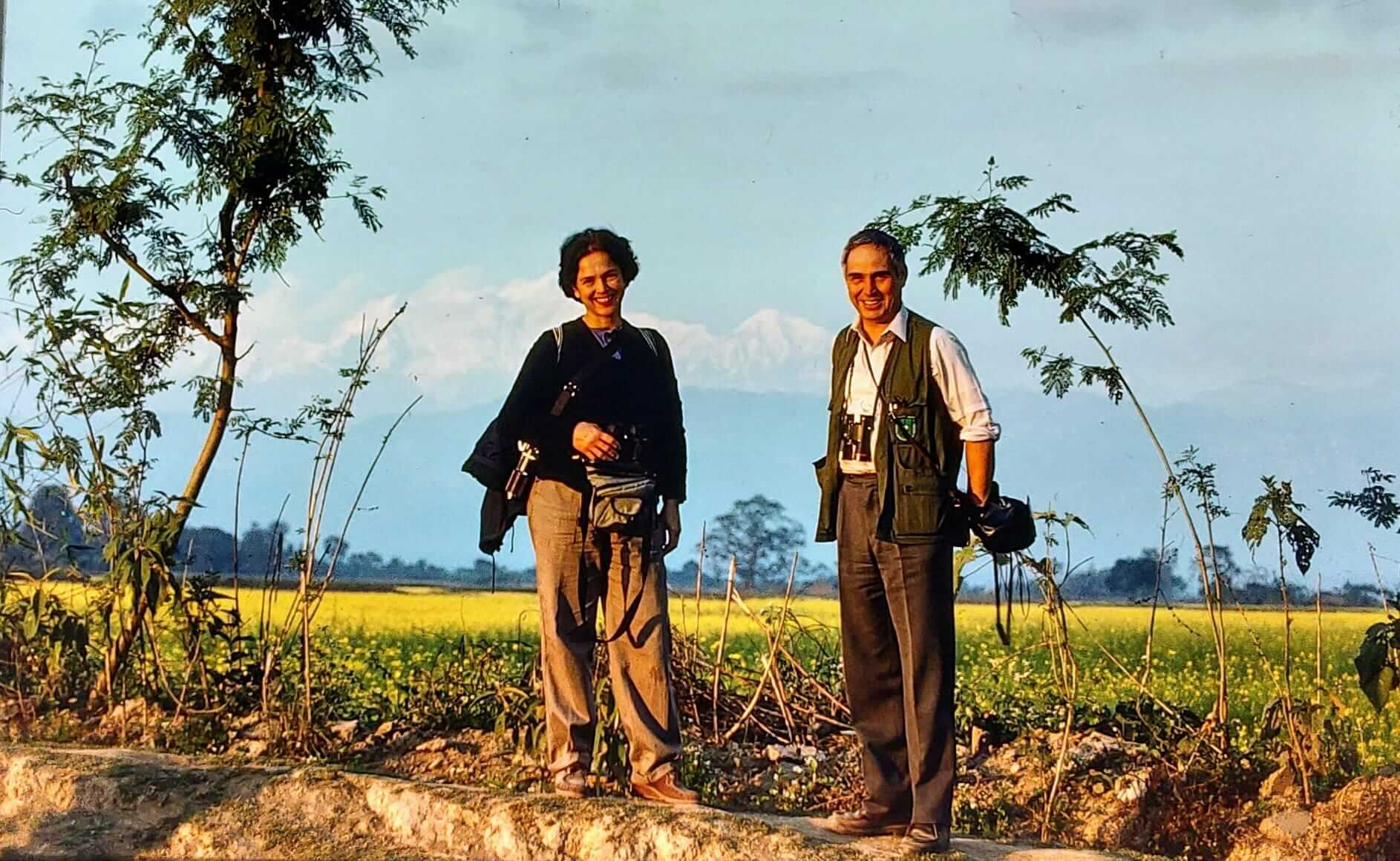 Due fotografi sorridenti posano in un paesaggio naturale, con montagne sullo sfondo e attrezzatura fotografica a disposizione.
