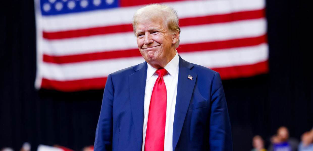 Trump sorridente durante un evento, mentre sullo sfondo sventola la bandiera americana, simbolo delle elezioni Usa e della crisi climatica.