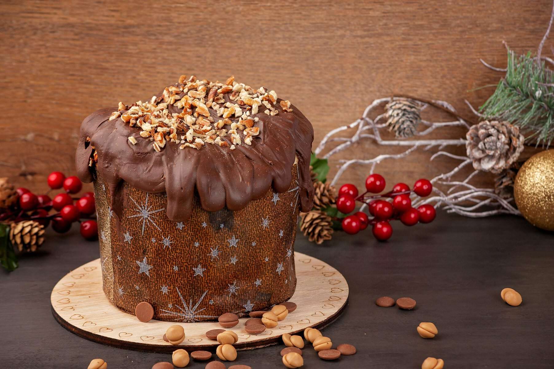 Panettone artigianale ricoperto di cioccolato e noci, decorato con bacche festive, ideale per le feste natalizie e ricco di proteine.