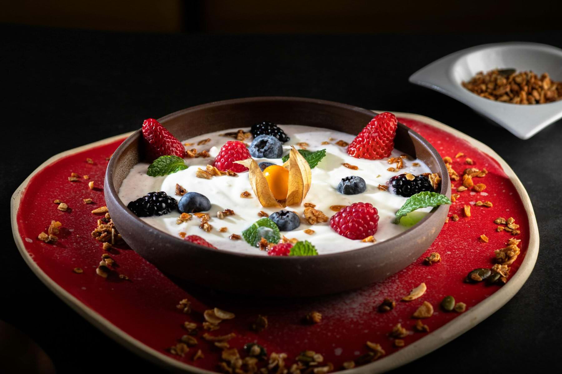 Ciotola di yogurt decorata con frutta fresca e granola su un piatto rosso, ideale per una dieta sana e preventiva contro il cancro al colon.