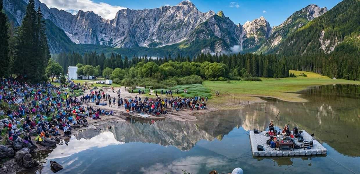 Il No Borders Music Festival compromette un'area protetta, la denuncia degli ambientalisti Concerto No Borders Music Festival in area protetta. Spettacolo musicale in un lago alpino con pubblico numeroso e maestose montagne sullo sfondo. Controversia ambientale.