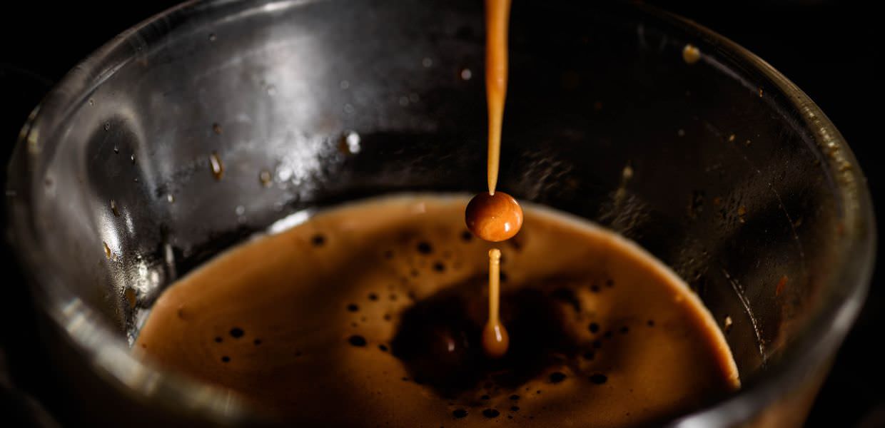 Ecco una descrizione alternativa SEO-friendly per l'immagine:

`Caffè espresso: goccia che cade in una tazza di vetro. Aumento prezzo caffè al bar: +103%. Crisi climatica tra le cause.`