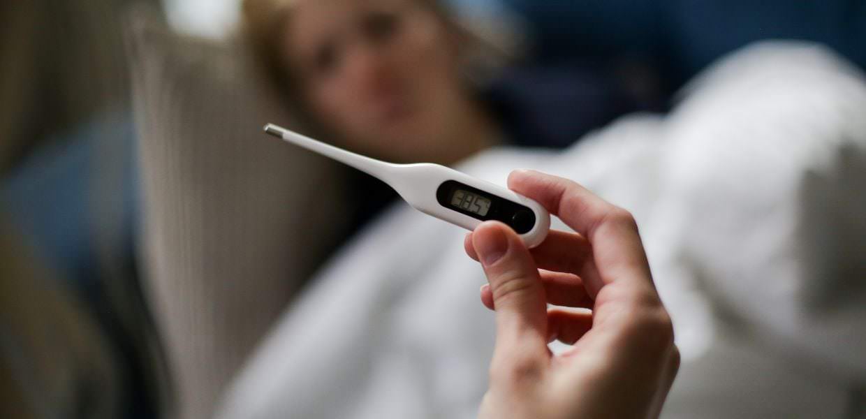 Un termometro digitale mostra una temperatura elevata, mentre una persona malata è visibile sullo sfondo, evidenziando l'aumento dei casi di influenza.